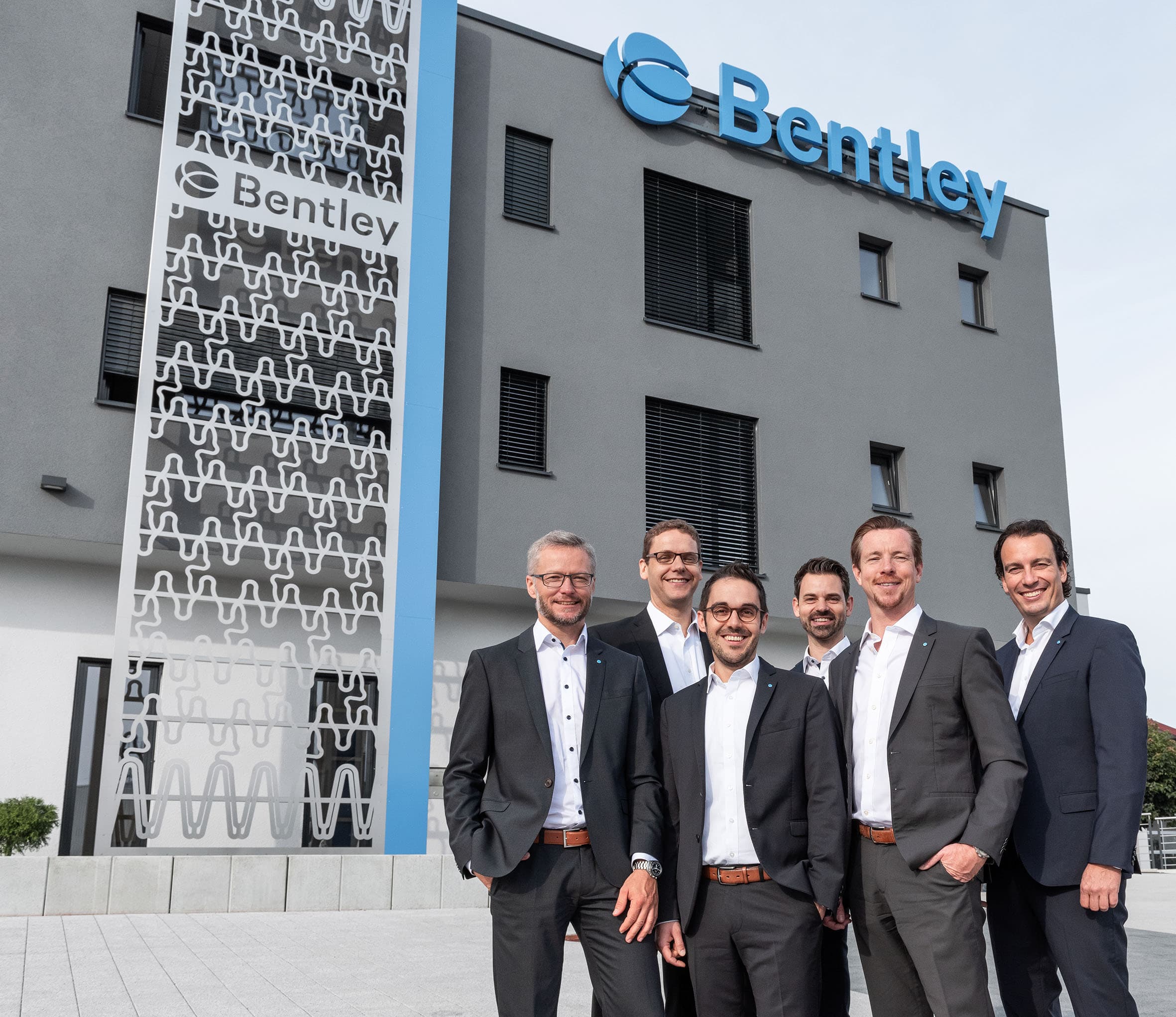 Six men in business attire stand smiling in front of a modern building with a "Bentley" sign and decorative facade.