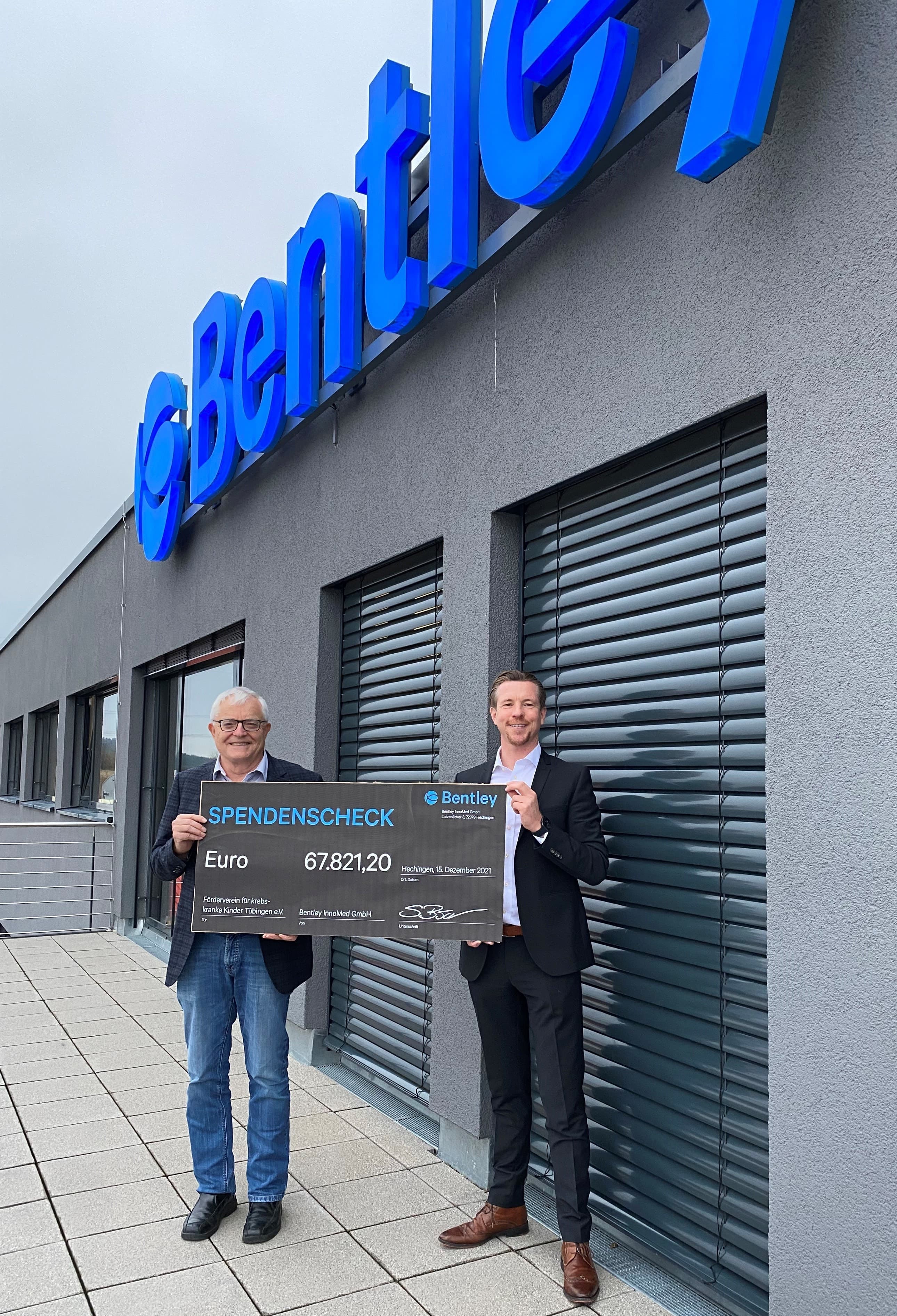 Two men holding a large donation check for 67,821.20 euros outside a Bentley building.