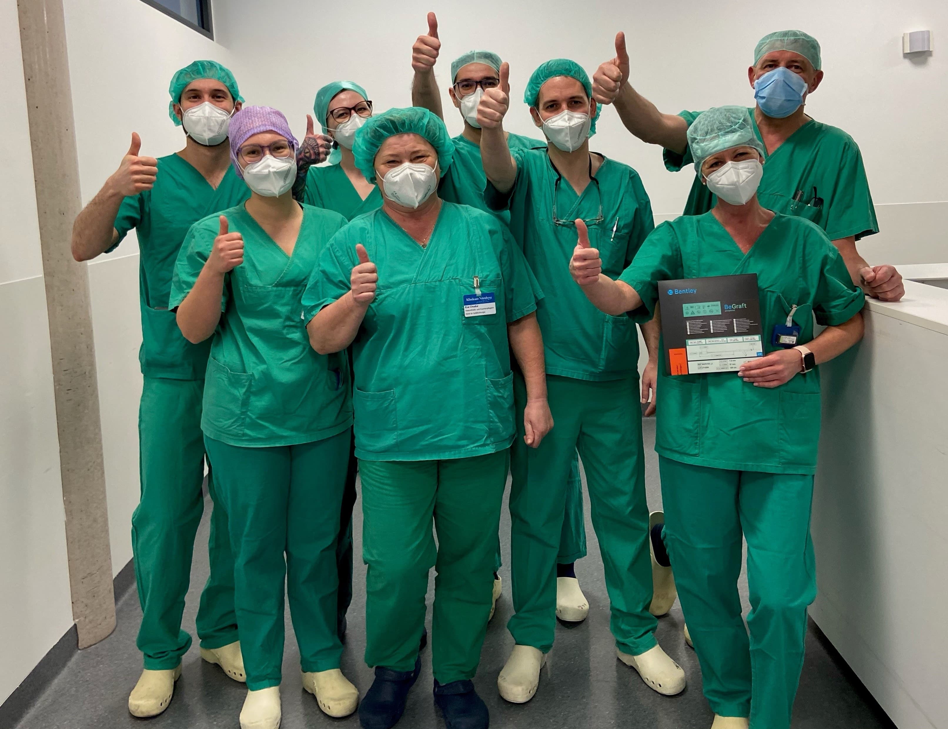 A group of medical staff in green scrubs and masks giving thumbs up in a hospital setting.