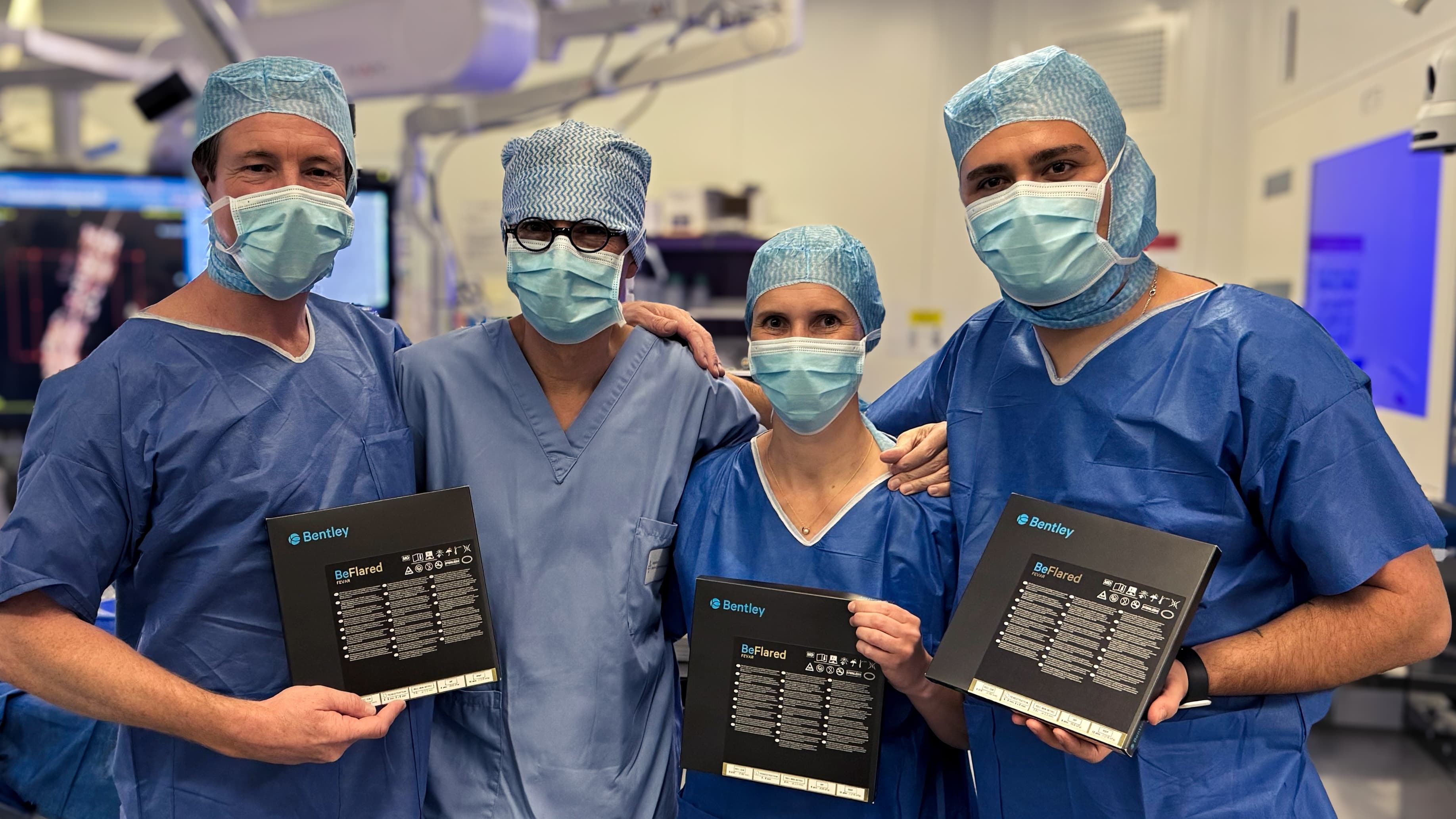 Four medical professionals in scrubs and masks hold Bentley certificates in an operating room.