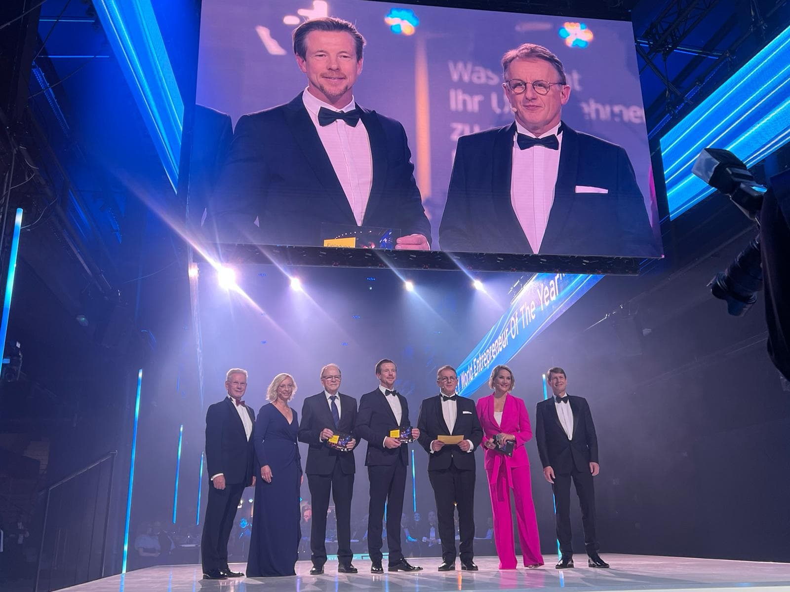 Seven people in formal attire stand on a stage under bright lights, with two men displayed on a large screen above them.