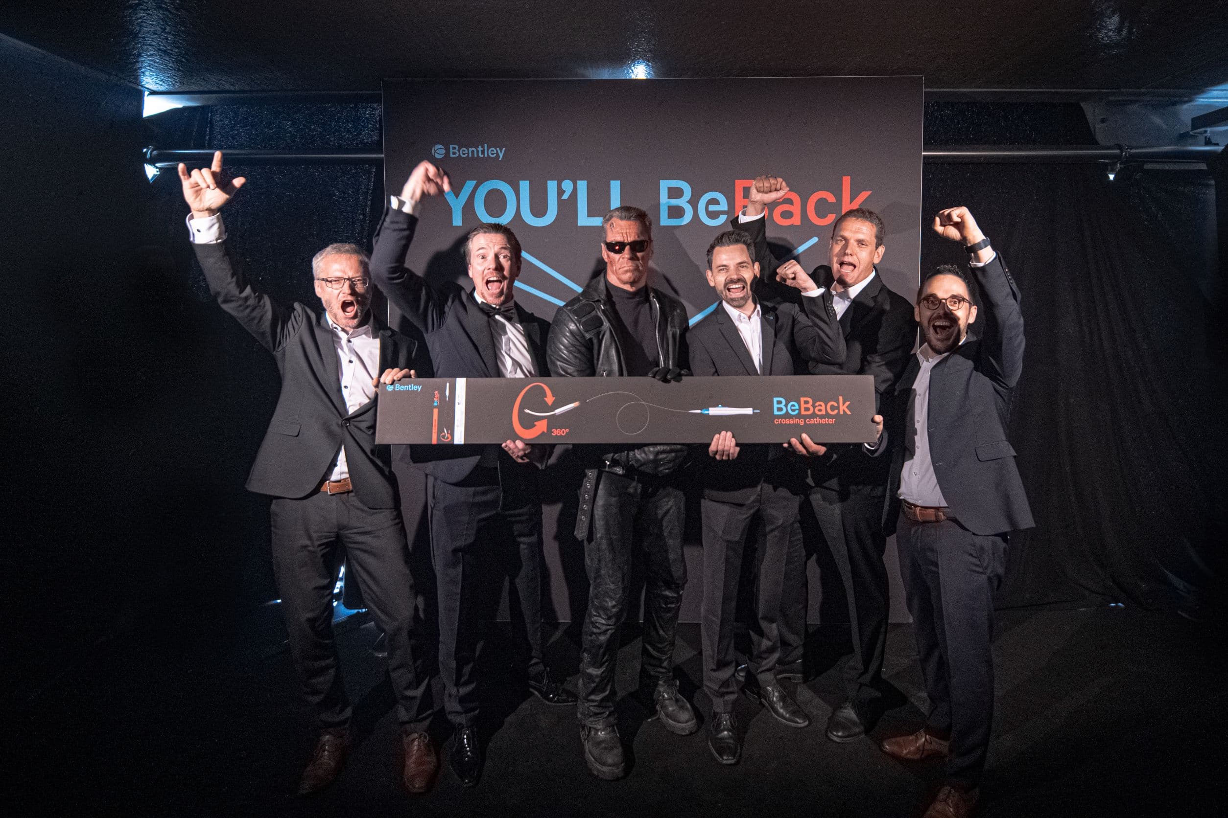 A group of six men in suits celebrate on stage, holding a large key with "BeBack" written on it, under a sign that says "YOU'LL Be Back."