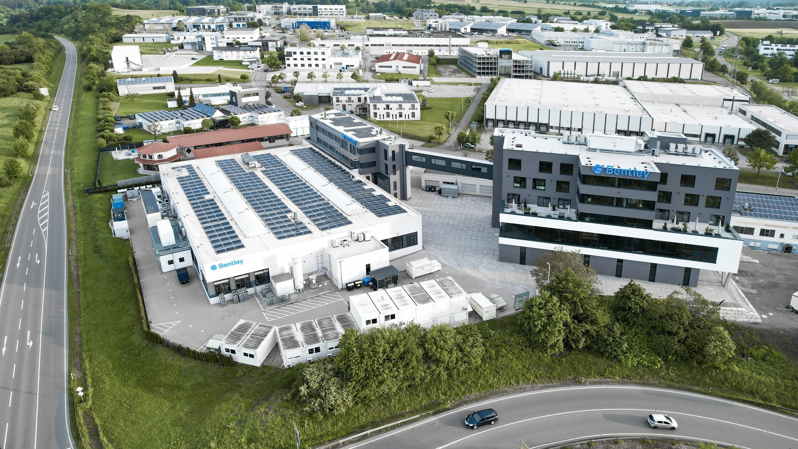 Aerial view of an industrial complex with solar panels on rooftops, surrounded by greenery and roads with vehicles.