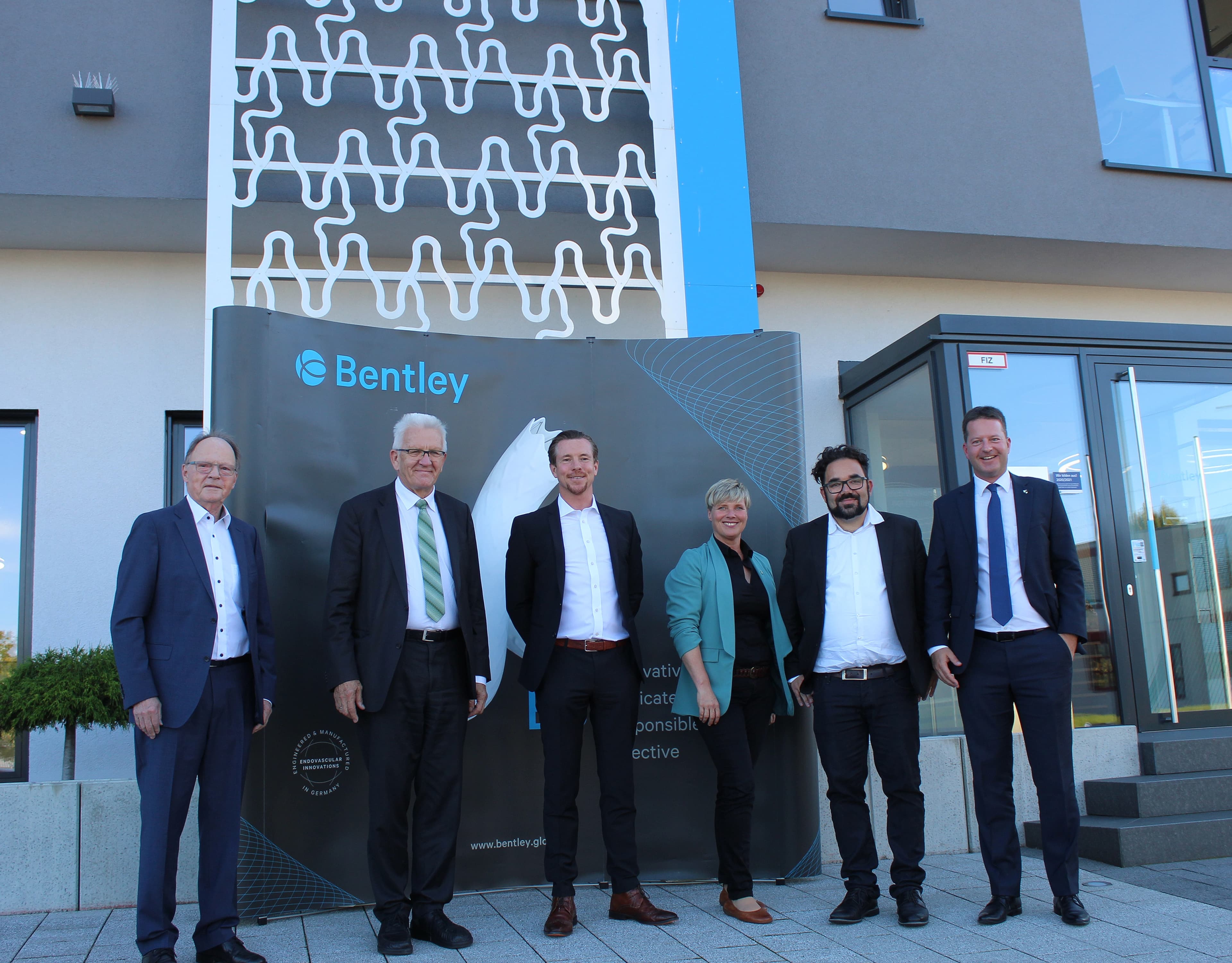 Six people in business attire stand in front of the Bentley sign and a modern building, posing for a group photo.